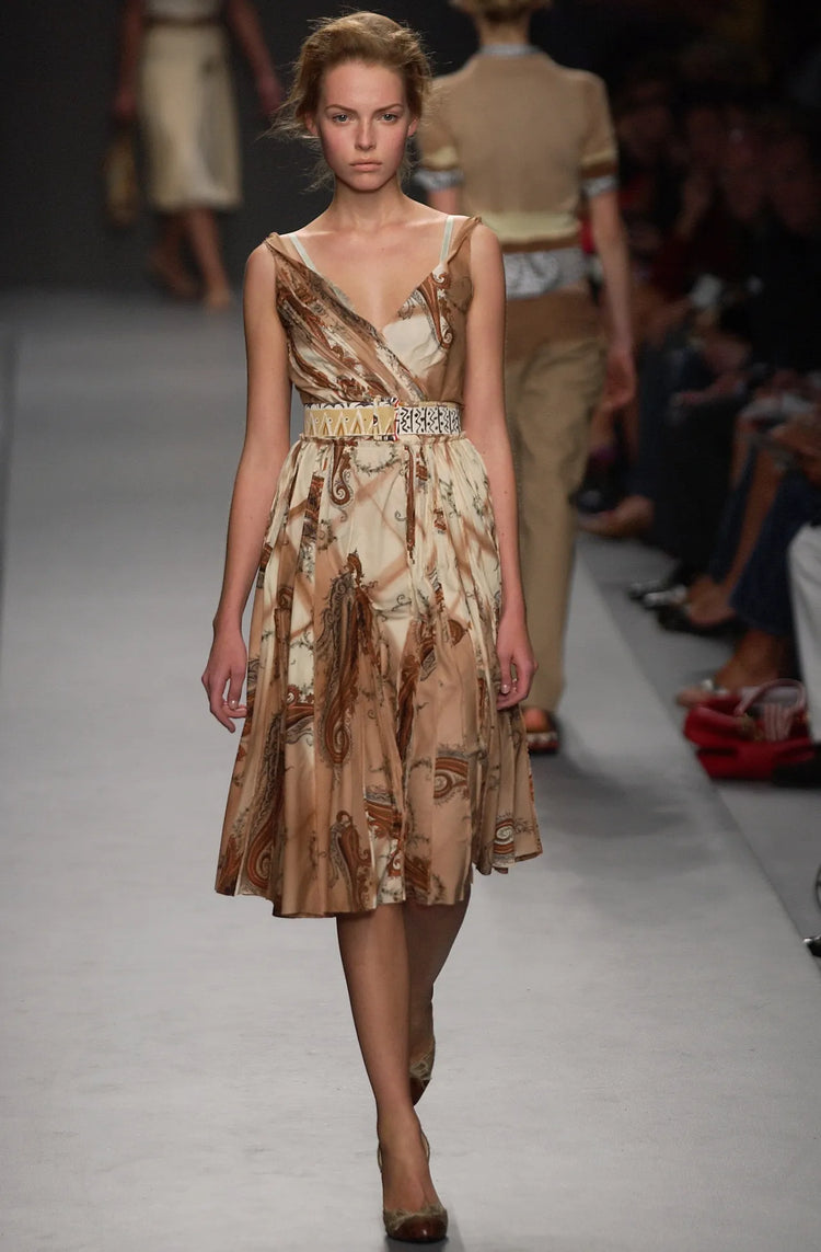Model walking on a runway wearing a patterned dress with a belt.