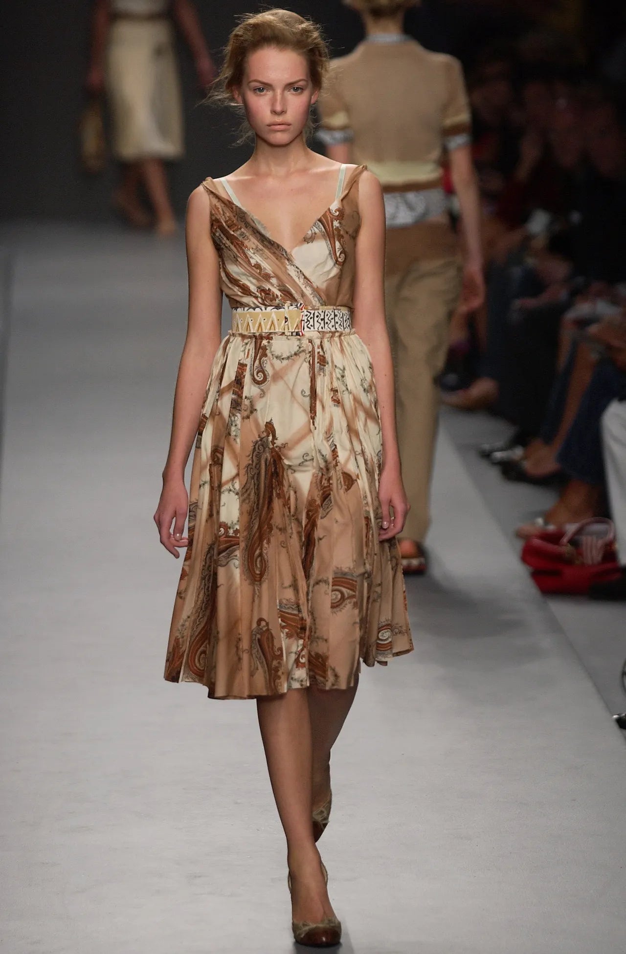 Model walking on a runway wearing a patterned dress with a belt.