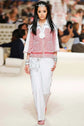 A model walks the runway wearing a Chanel fashion show outfit featuring a red and white tweed vest with silver trim, a white collared shirt, white trousers with red piping, and a matching red and white tweed clutch bag.