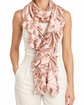 A woman wearing a long, ruffled scarf in a soft blush pink satin or silk-like fabric, styled over a white sleeveless top and white trousers against a plain white studio background.